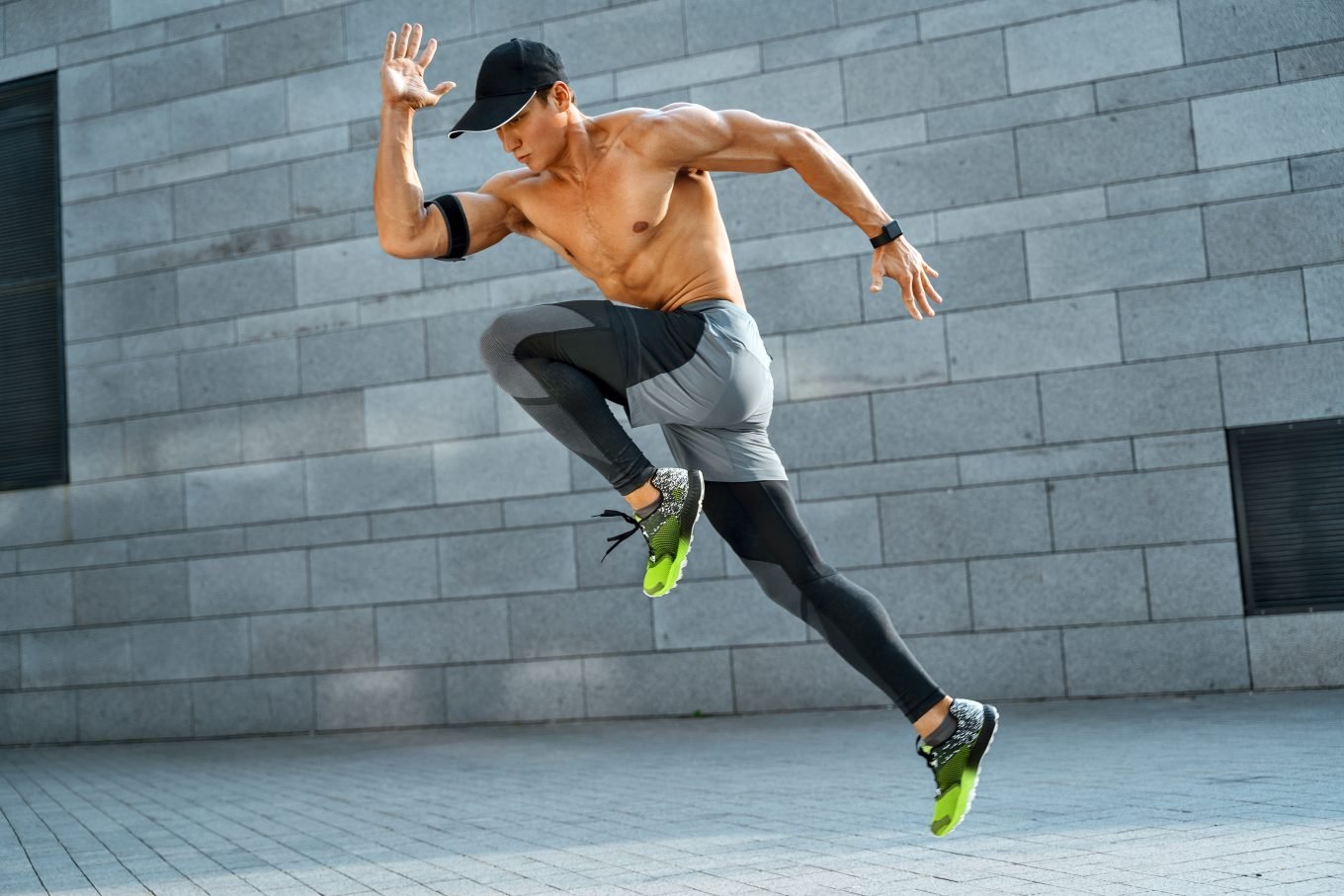 The photograph shows an athletic man in an energetic jump pose in an urban environment. One knee is drawn up to his chest and the other extends behind him. His arms are positioned as though running. He is wearing sports leggings, trainers, a cap, a heartrate monitor on his arm, and a sports watch. The photo intends to demonstrate the power of fitness testing for athletic performance. 