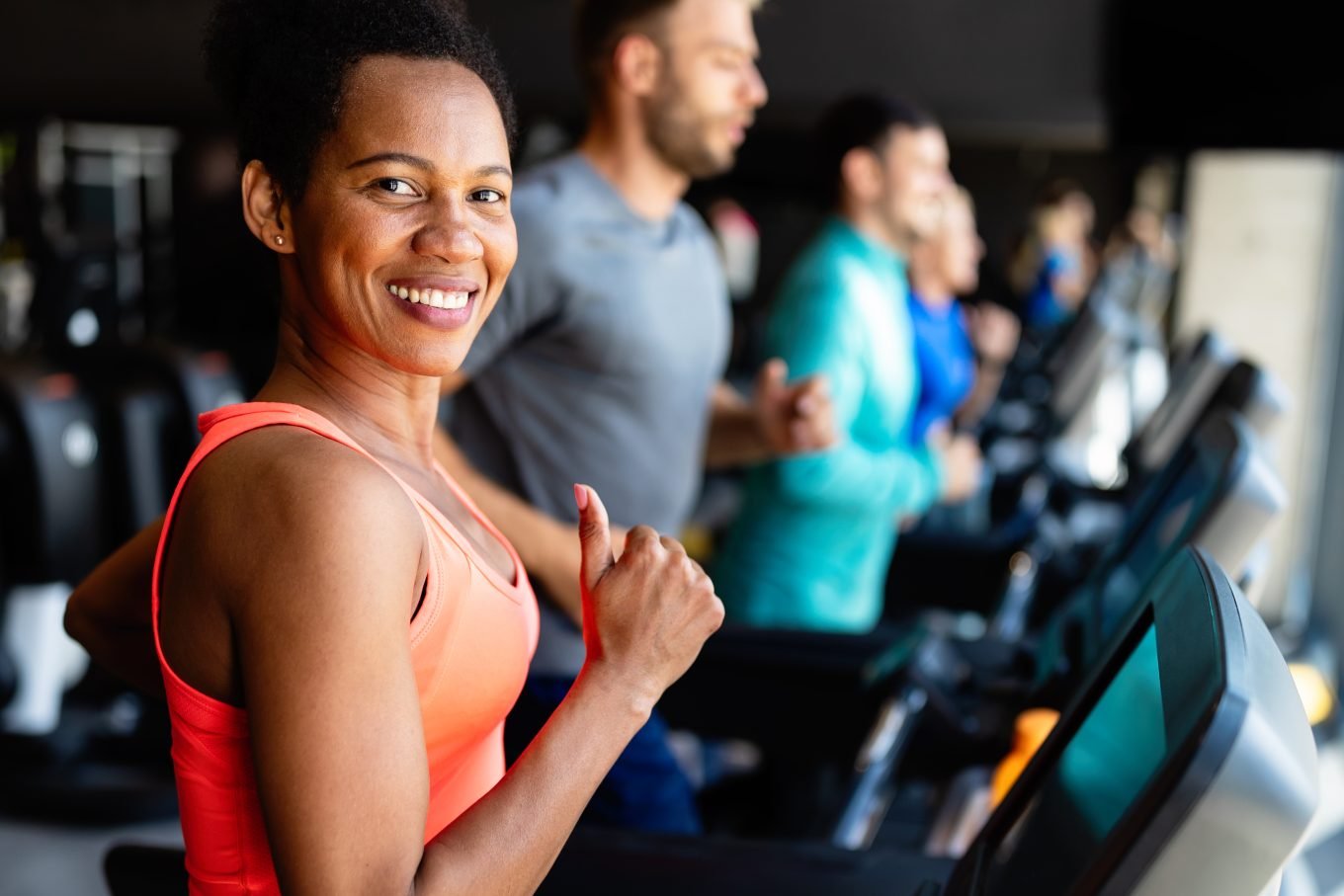 The image shows an athletic black woman making a thumbs up signal while working out on a treadmill at the gym. There are several other gym-goers visible on treadmills in the background. 