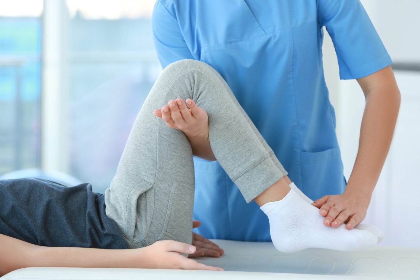 The photograph is a cropped view of a client lying flat on their back with one knee bent. A physiotherapist or functional movement assessor has one hand supporting the raised knee, and the other hand gently pressing down on the corresponding foot. The image is intended to demonstrate a functional movement assessment in action. 