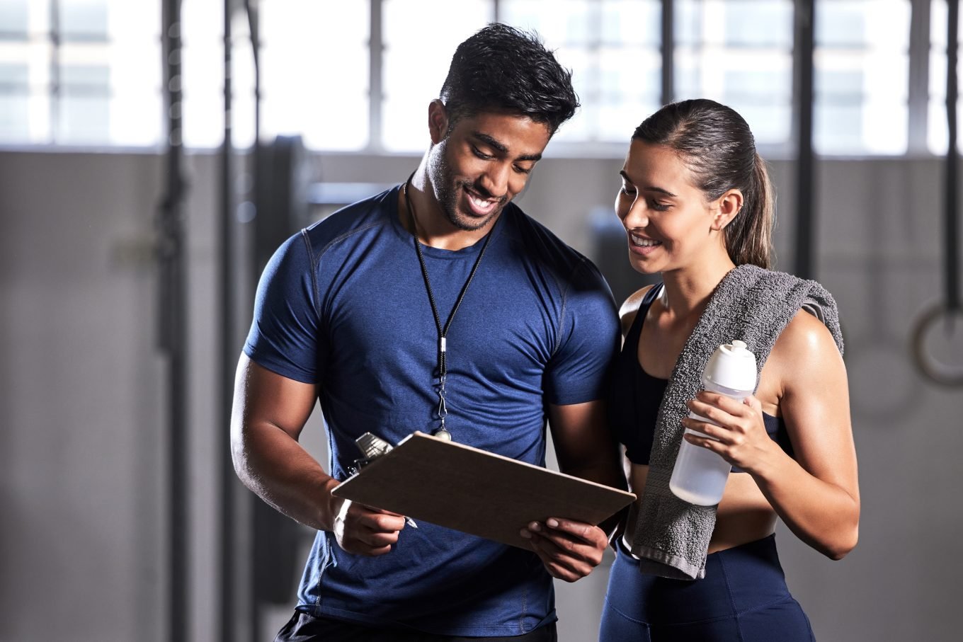 A brown, male personal trainer consults a clipboard with test results during a training session with a white, female client, who is holding a water bottle. The photo aims to illustrate the benefits and limitations of fitness testing in a personal training context.
