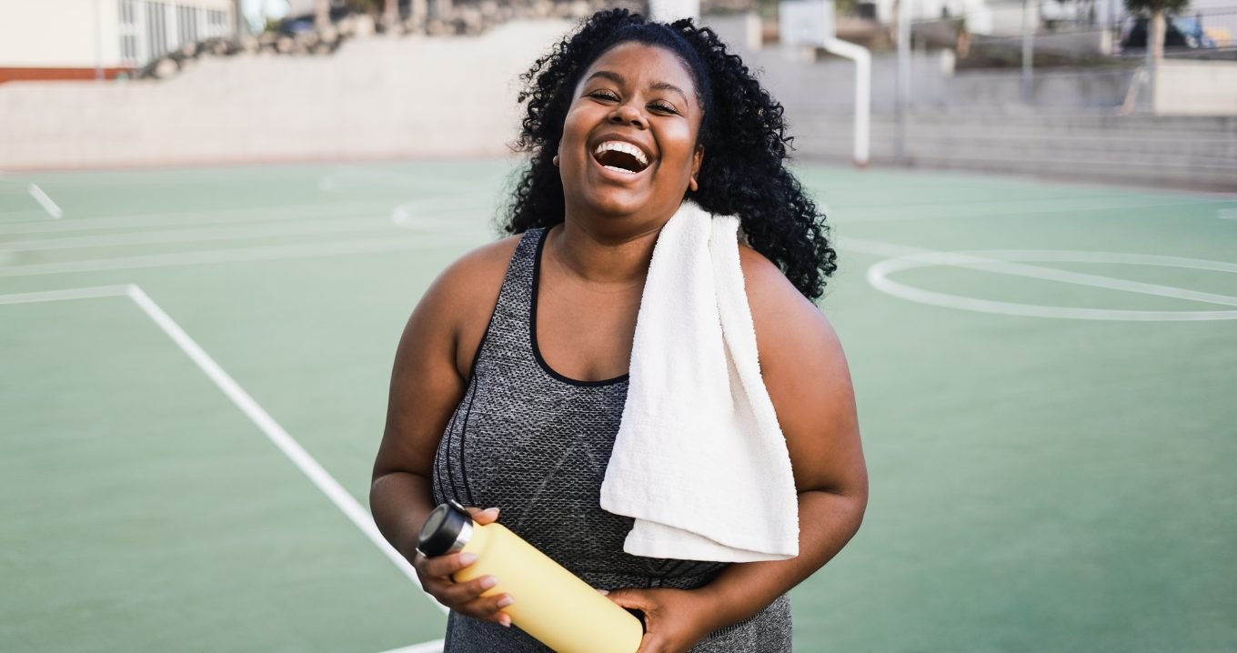 A black woman smiles while standing on a green basketball court. She is dressed in a grey workout top, is holding a yellow water bottle, and has a white workout towel over her shoulder. 