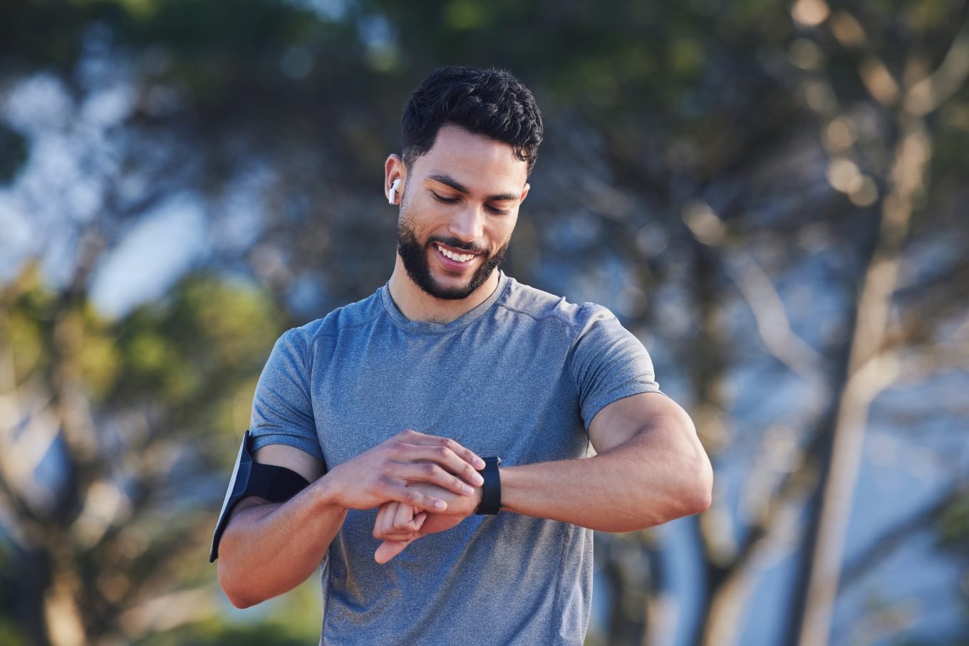 A brown-skinned man with black hair and a beard, wearing a workout shirt, a phone-holder armband, and wireless headphones checks his fitness watch while running in the park. Trees and a blue sky are visible in the background. 