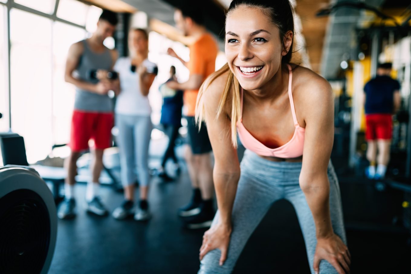 A woman stands resting her hands on her knees, grinning, in an exercise studio. She is dressed in grey workout leggings and a pink workout top, with her hair tied in a ponytail. Other athletes are visible in the background. 