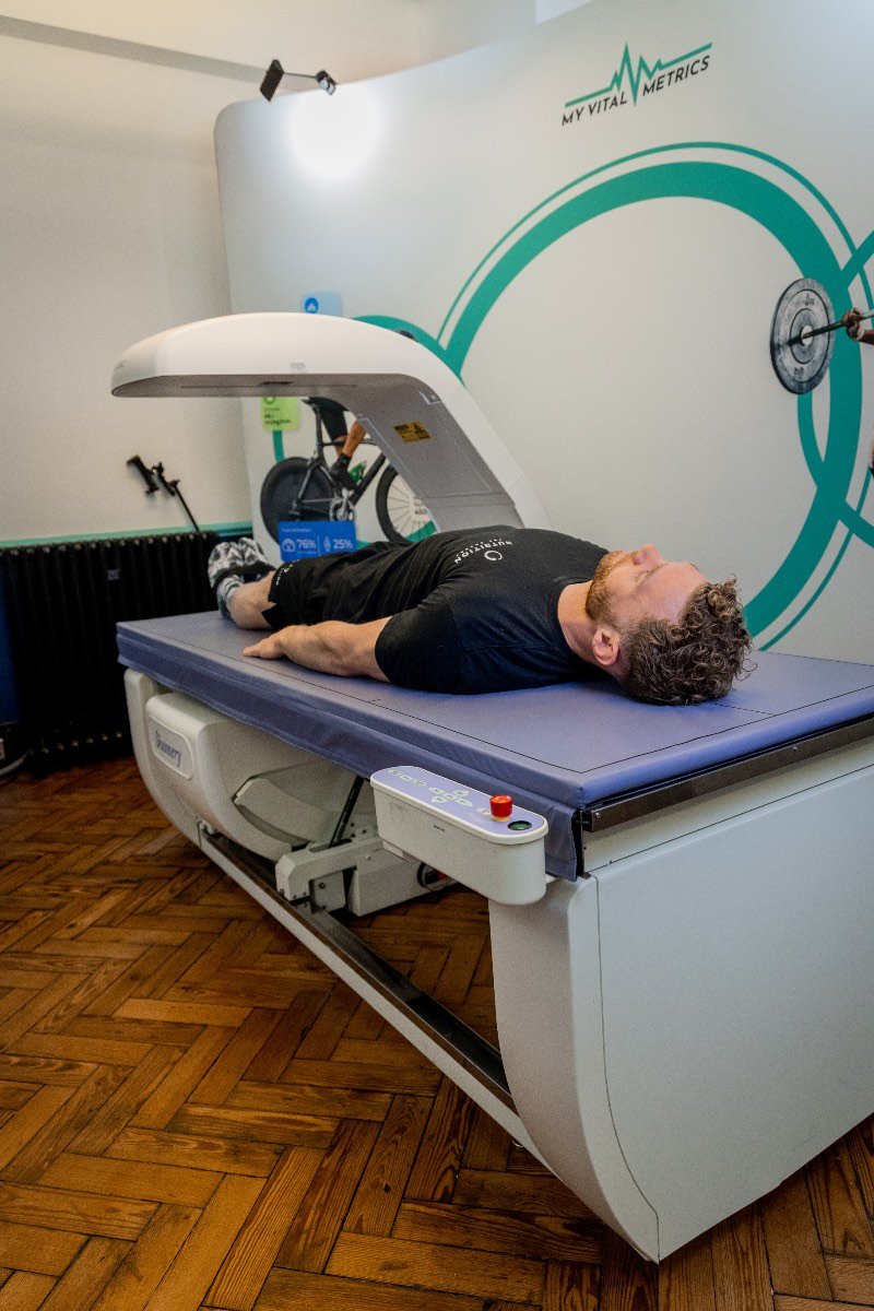 The photo shows a white man having a body composition scan in a DEXA scanner. He is lying flat on his back on a raised table as the arm of the scanner passes over his body. There is a logo on the wall behind him that reads My Vital Metrics.