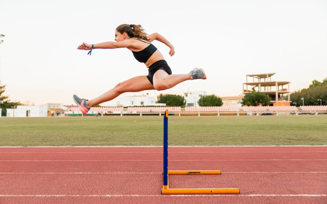 A female athlete clears a hurdle on a red, outdoor running track. She is suspended midair in a powerful jump, her arms propelling her forwards through the motion. She wears a triumphant expression.