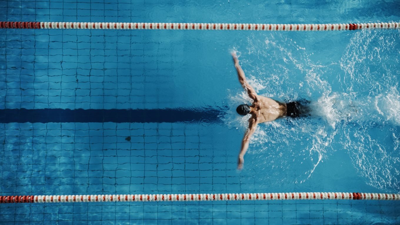 An aerial photo shows a muscular male swimmer doing breaststroke in a bright blue indoor swimming pool. The swimmer is wearing black swimming trunks and swims in a lane separated by two rows of floating red and white lane-dividers. He swims in line with the black line on the bottom of the swimming pool. 