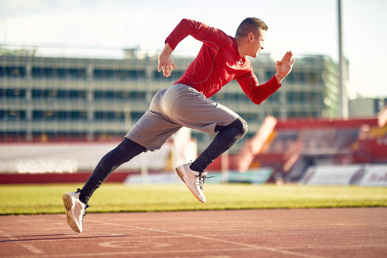 A male athlete completes a powerful sprint drill on a red, outdoor running track. He is suspended midair by the power of his run. He is wearing a red athletic shirt, grey athletic shorts, and black running tights. 