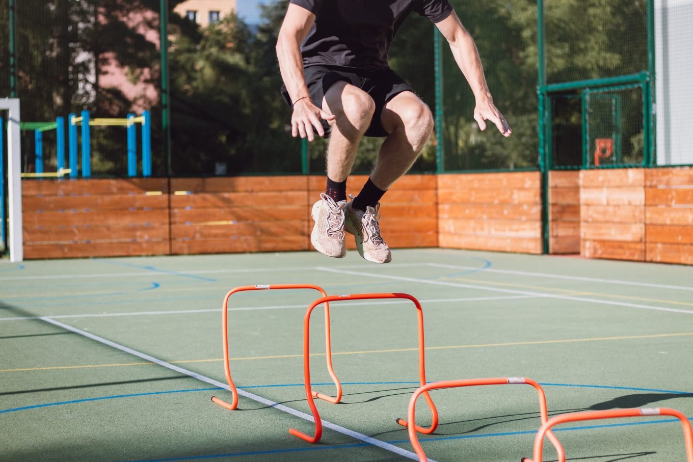 A white, male athlete dressed in black jumps with feet together over a plyometric hurdle, as part of an agility hurdle drill. he is on a basketball court, and trees and a fence are visible in the background. 
