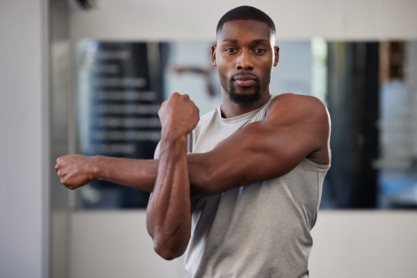 A muscular black man with a cropped haircut and wearing a grey tank top is visible from the waist up in an indoor setting. He has one arm stretched across his body and is using the other arm to stretch his upper body. 