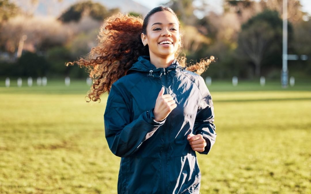A brown-skinned woman runs in a field. She is dressed in a navy athletic jacket and her curly hair is tied in a ponytail and flows behind her as she runs. She is smiling and enjoying the run.