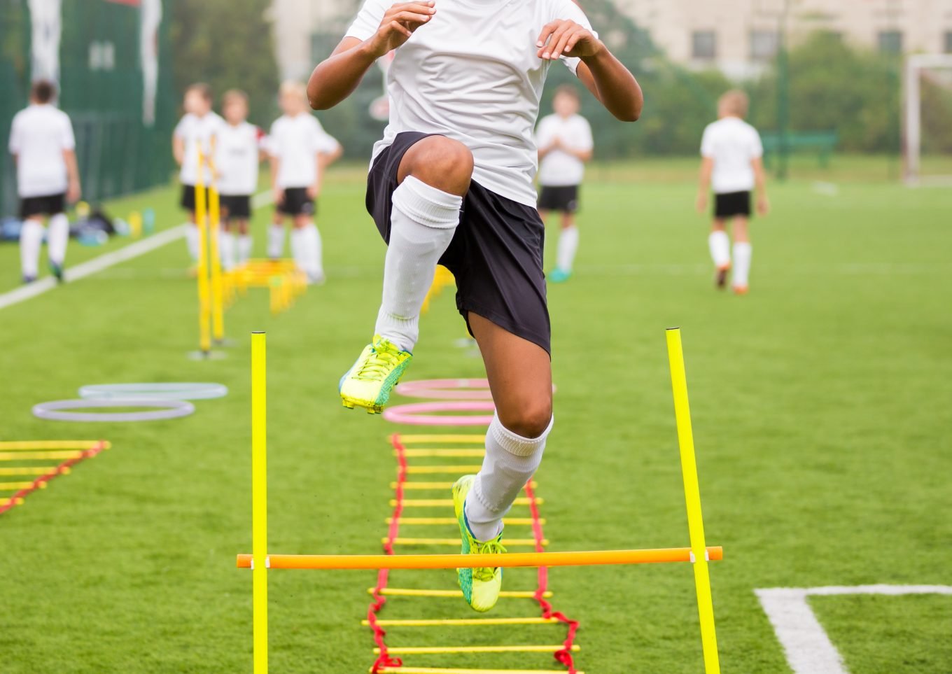 A young male athlete jumps a plyometric hurdle after having completed an agility ladder drill. Other althetes are visible in the background on a grassy football field. 