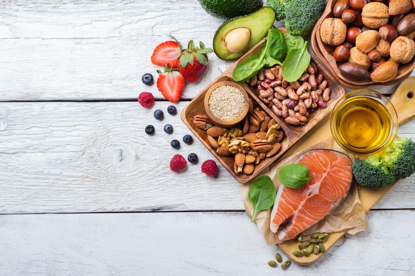 An array of raw healthy food items are displayed on a wooden platter atop grey wooden floorboards.  A salmon filet, an avocado, basil leaves, and various nuts and berries are visible. 