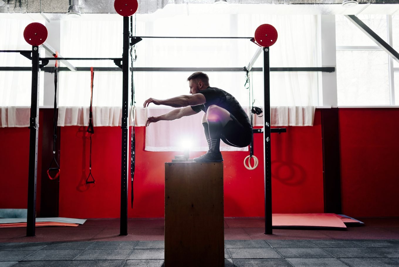 A white, male athlete lands a plyometric box jump inside a gym. He is crouched on top of a tall box having jumped up as part of an agility drill. Fitness equipment is visible in the background against a red wall. 