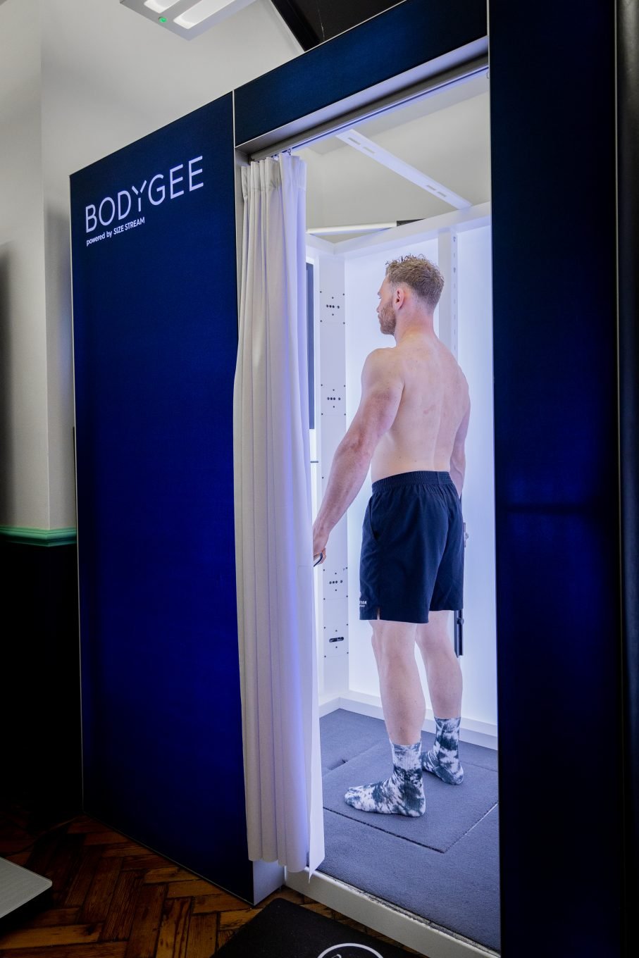 The photo shows a white man having a body composition scan in a DEXA scanner. He is lying flat on his back on a raised table as the arm of the scanner passes over his body. There is a logo on the wall behind him that reads My Vital Metrics.