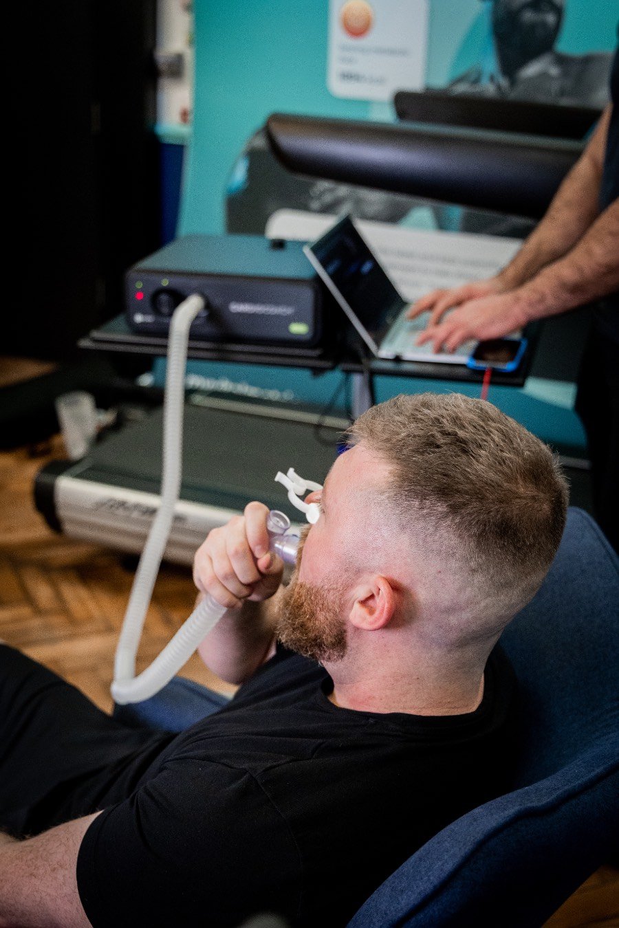 The photo shows a white man having a body composition scan in a DEXA scanner. He is lying flat on his back on a raised table as the arm of the scanner passes over his body. There is a logo on the wall behind him that reads My Vital Metrics.