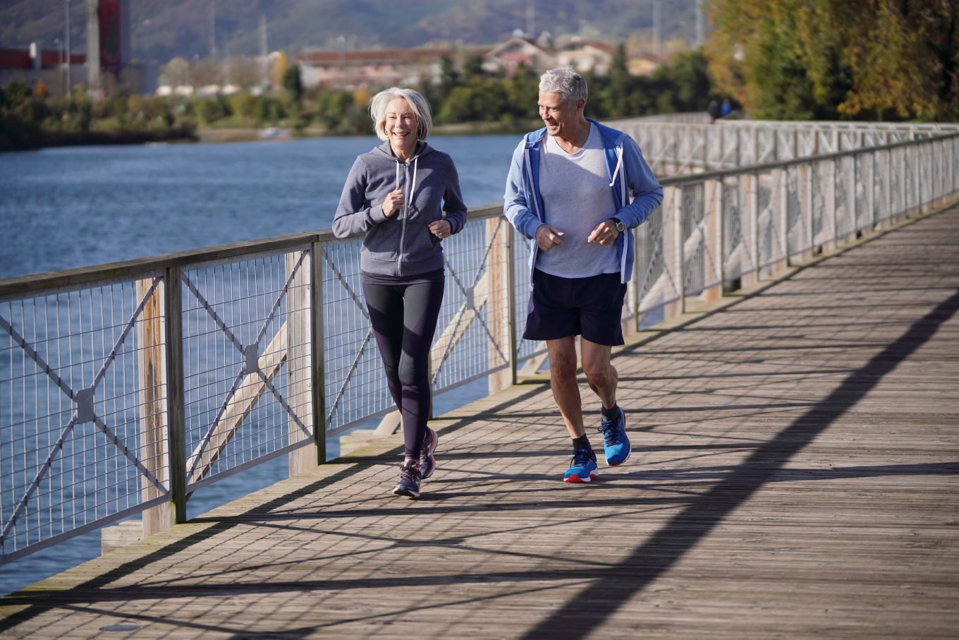 An older man and woman run together alongside a river. The sun is shining and a metal barrier separates the path from the water. Both runners are wearing workout clothing, and are smiling as they complete the workout. 