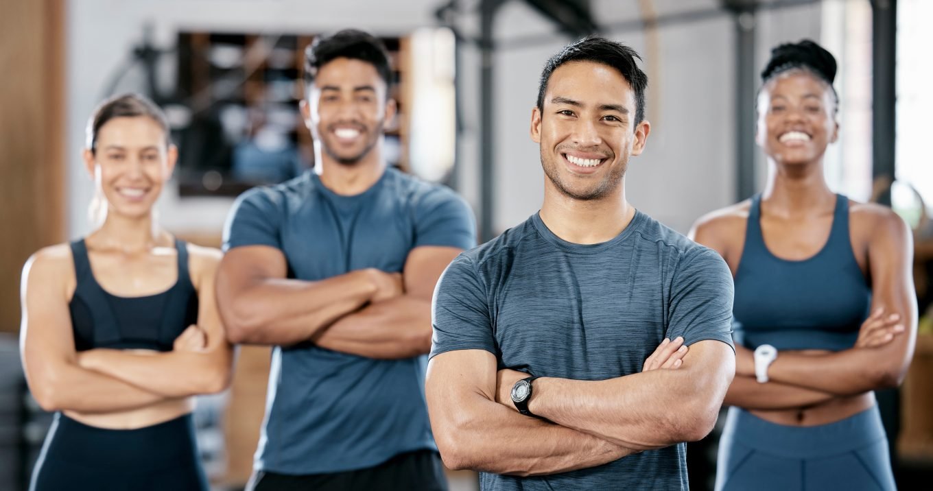 A team of personal trainers of a variety of genders and racial backgrounds stand with their arms crossed, grinning, in front of a blurred gym background.  