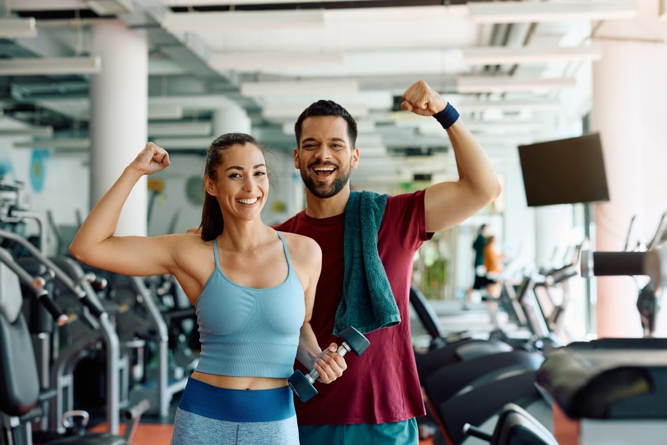 A man and a woman stand triumphantly in a gym, each smiling and posed to flex a bicep. They are holding a dumbbell and a gym towel. 