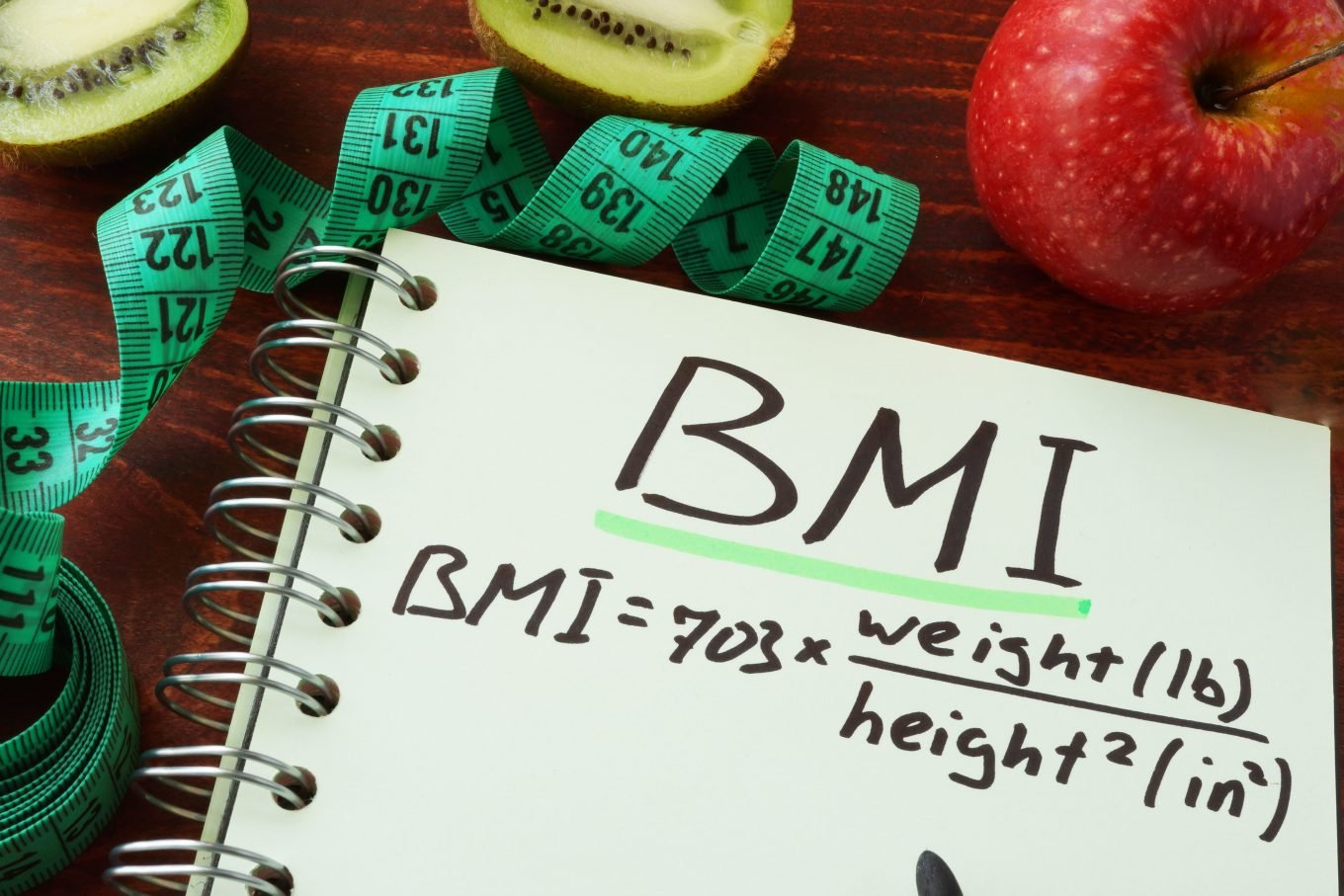 An open notebook, in which the formula for calculating BMI is written, sits atop a table alongside a green, curled tape measure, half a kiwi, and a red apple. 