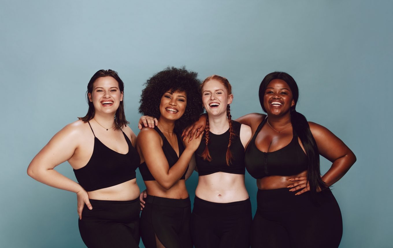 A group of four young women stand posed in gym clothing with their arms around each other, smiling and laughing. 
