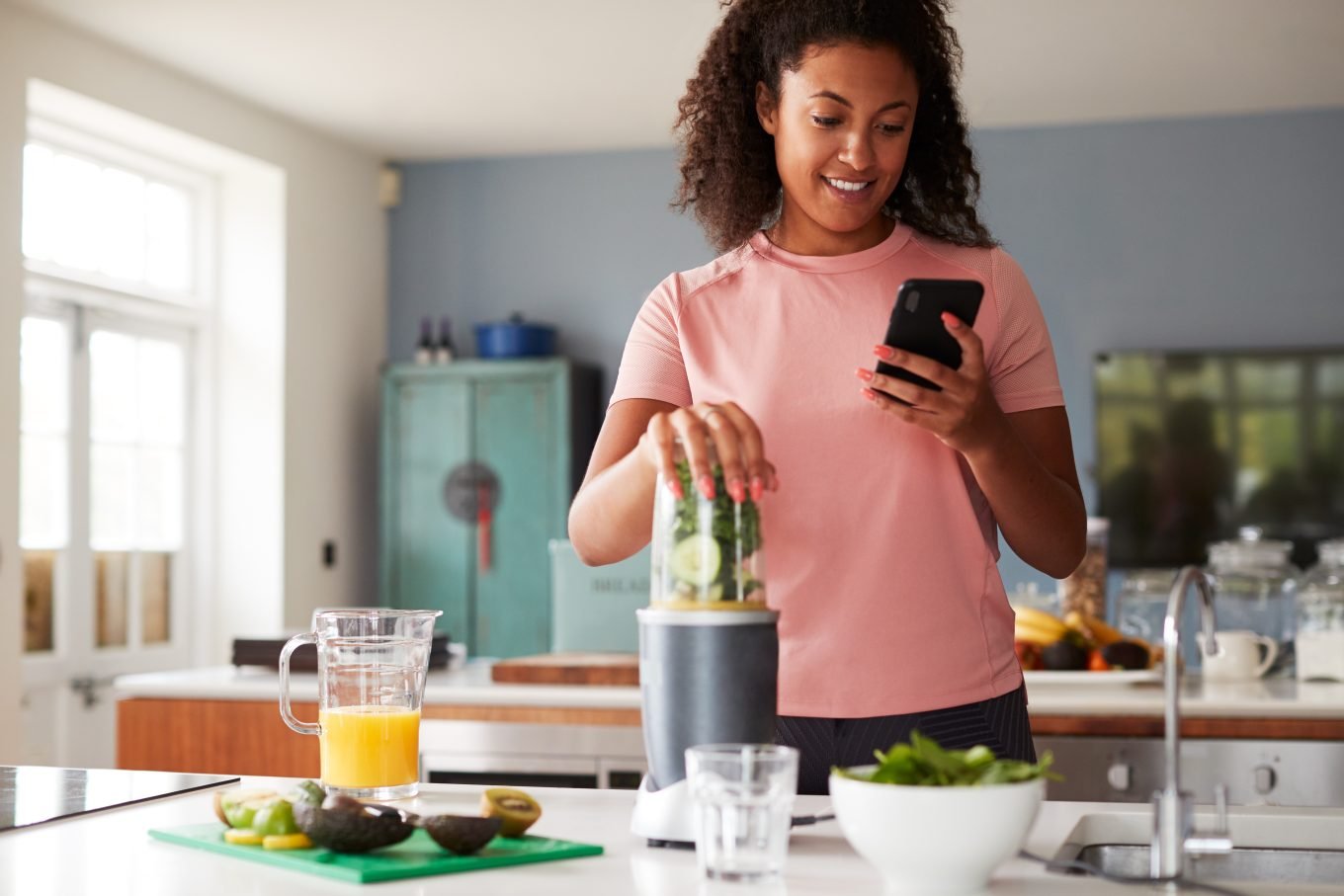 A dark-skinned, curly-haired woman looks at her phone while blending fruits and vegetables to create a healthy smoothie. 