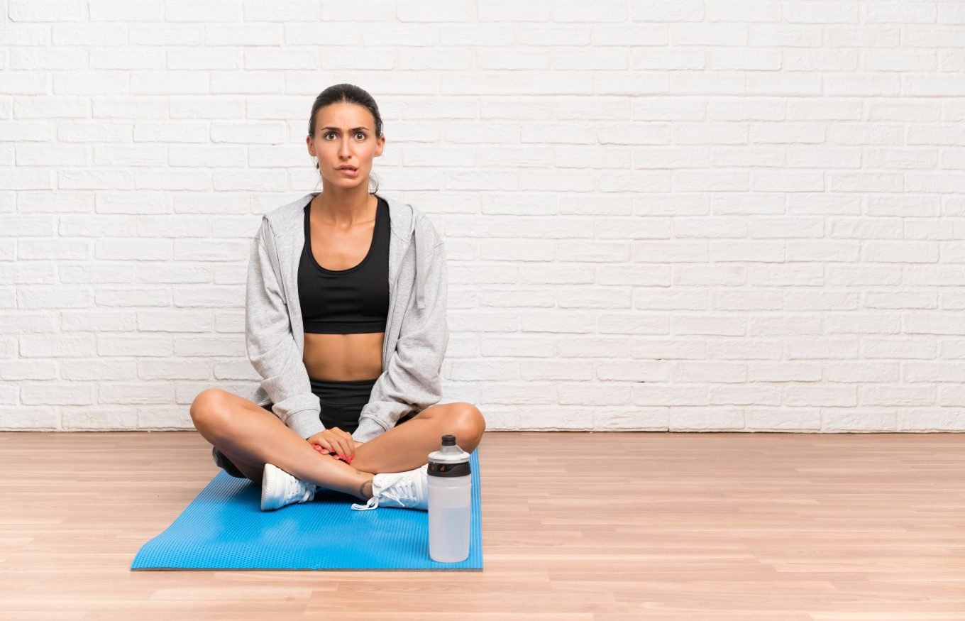 A young caucasian woman with a perplexed look on her face sits cross-legged on a blue yoga mat. She is wearing gym clothing and a water bottle sits in front of her. 