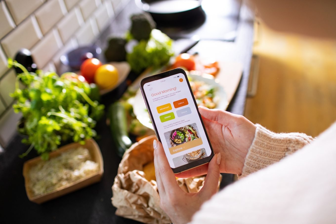 A phone screen displaying a calorie tracker app is held over a kitchen counter on which fresh fruit and vegetables are visible.