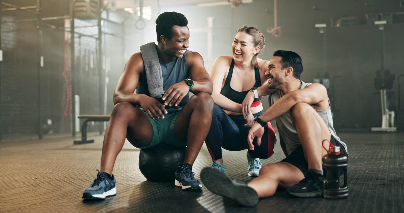 A group of three young adults are casually gathered in a gym, two crouching and one sitting. They are all wearing workout attire and smiling, perhaps sharing a joke or celebrating a win. 