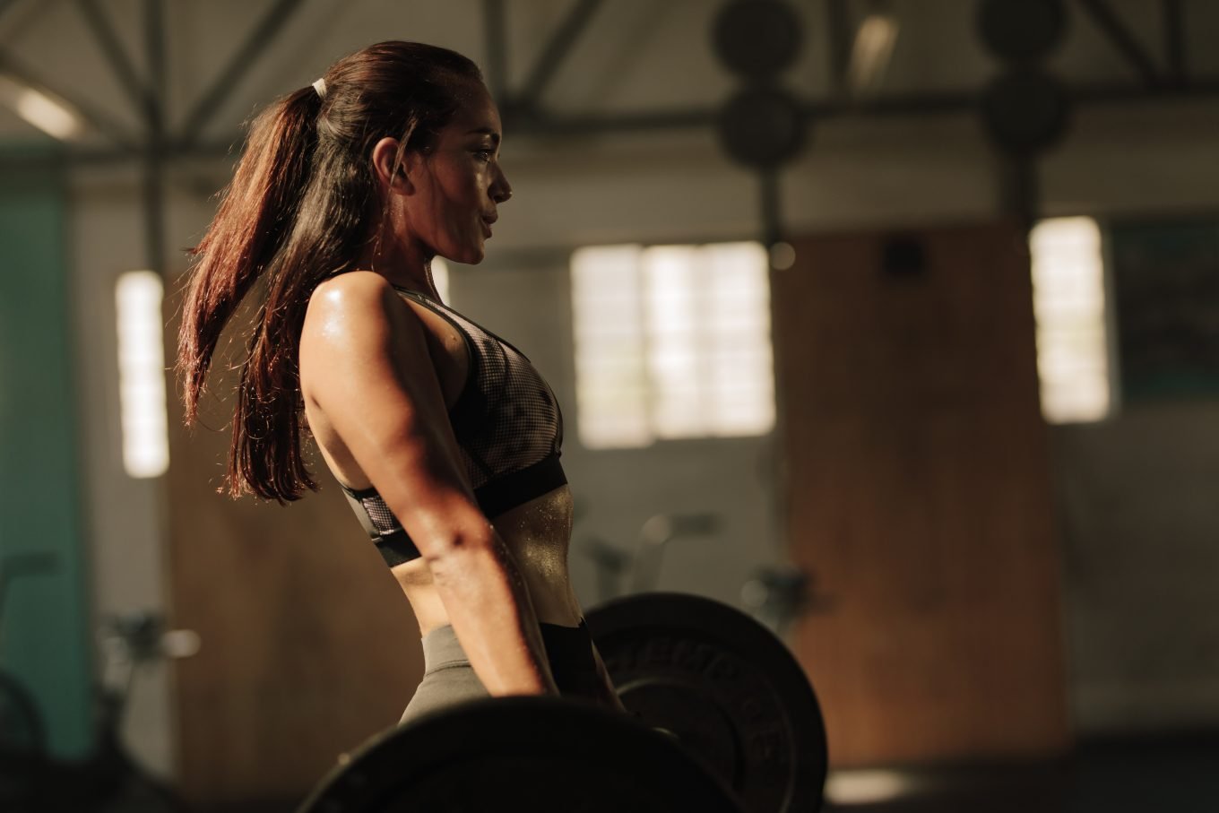 An athletic woman completes a heavy deadlift with a barbell in a gym with low lighting. 