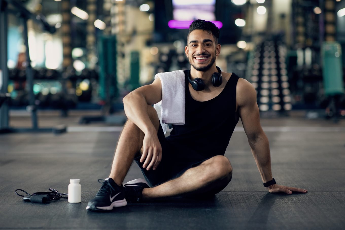 A young, athletic man sits smiling on the gym floor, with a white towel folded over his shoulder and headphones around his neck. Gym equipment is visible in the background, and a bottle and a resistance band are on the floor beside him. 