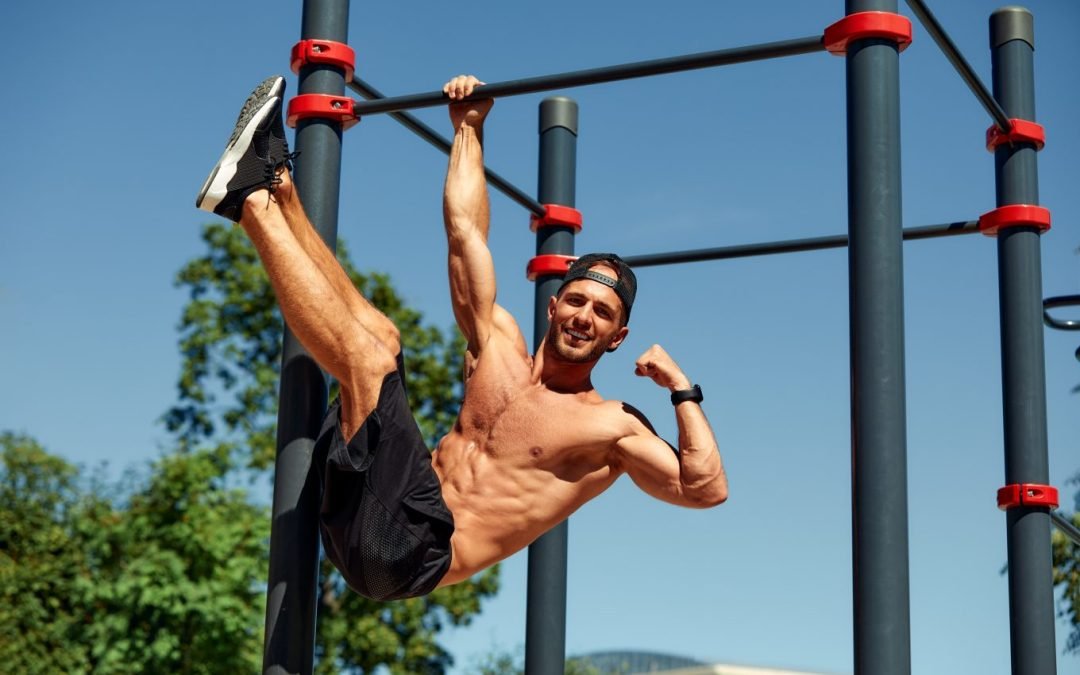 A muscular young man hangs from one arm from an outdoor climbing apparatus, grinning with his legs crunched upwards and the other bicep flexed.