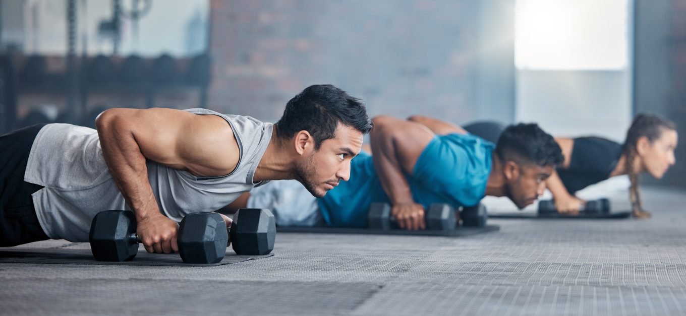 Two men and a woman lie on the floor ready to perform a push-up with their hands placed on dumbells. 