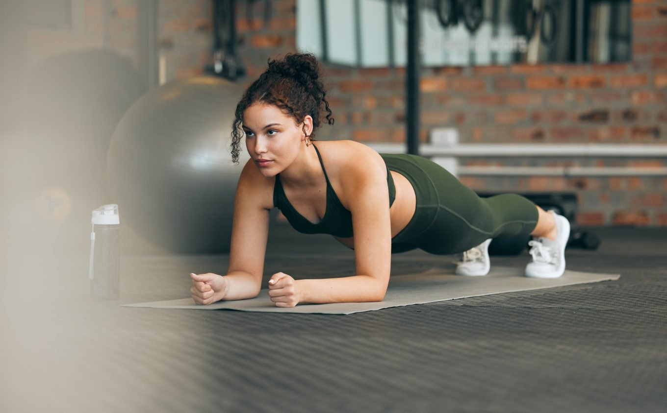 A young woman holds a plank position on a yoga mat in the gym. A gym ball and other gym equipment is visible in the background. 