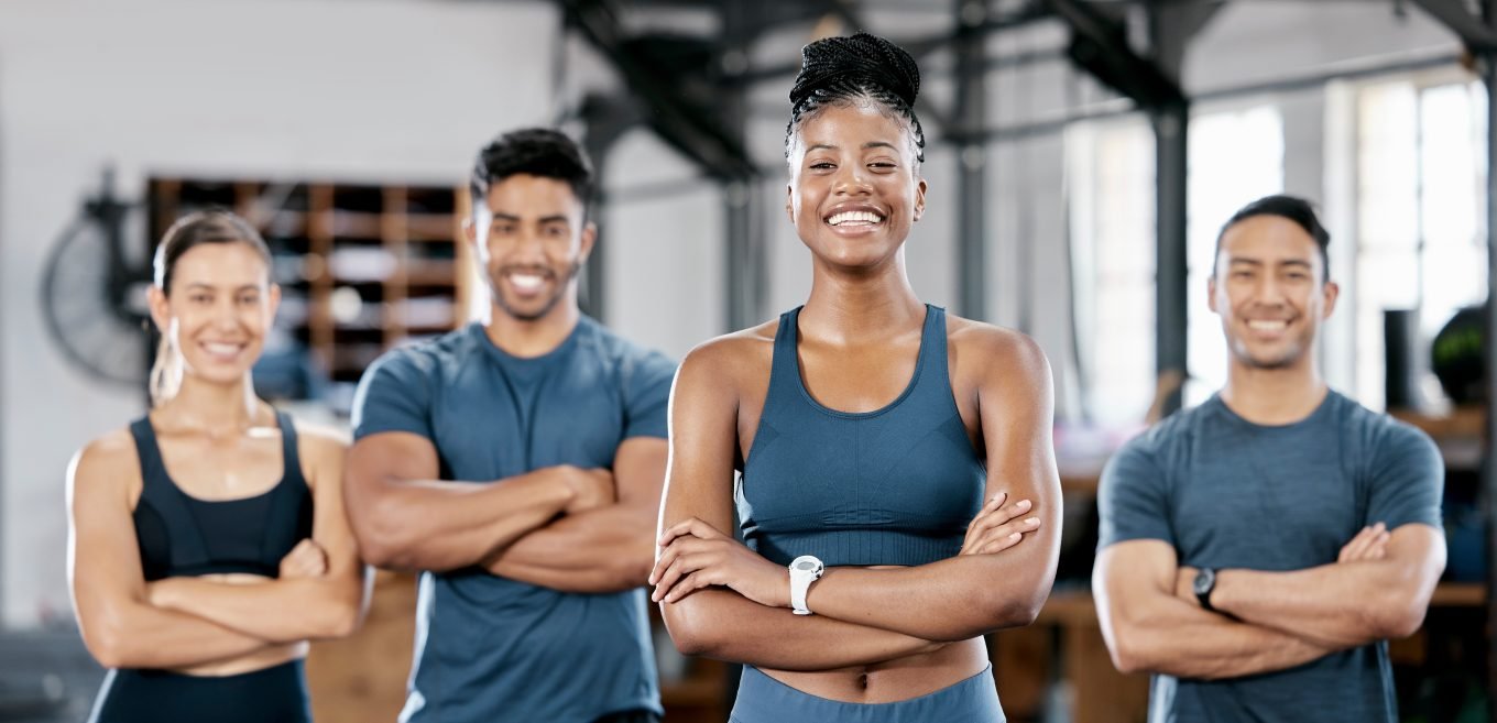 A multiracial team of 4 personal trainers in blue gym clothes smile with their arms crossed. 