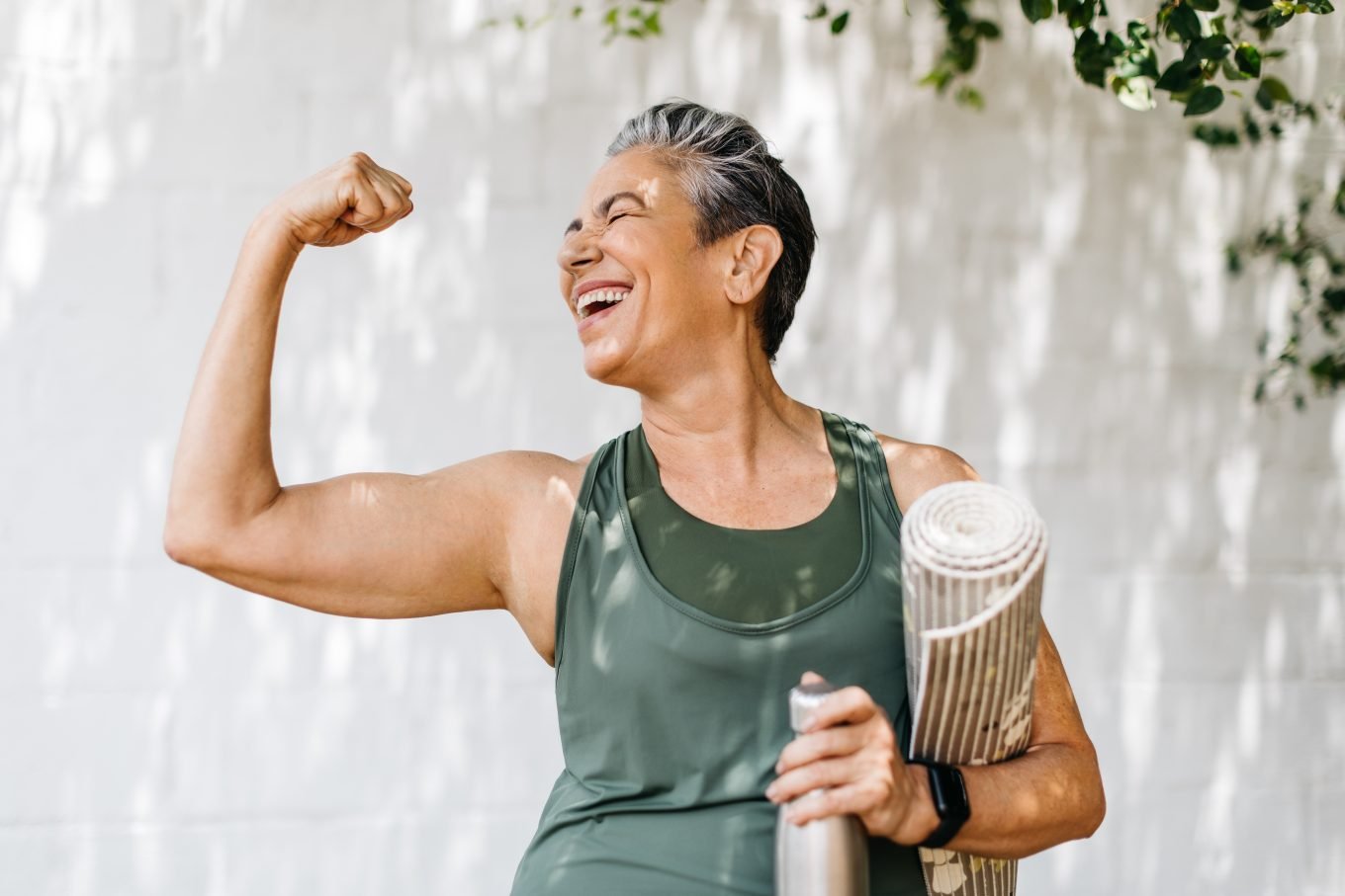 A mature woman in green workout clothing holds a yoga mat and water bottle under her left arm while flexing her right bicep, smiling. 