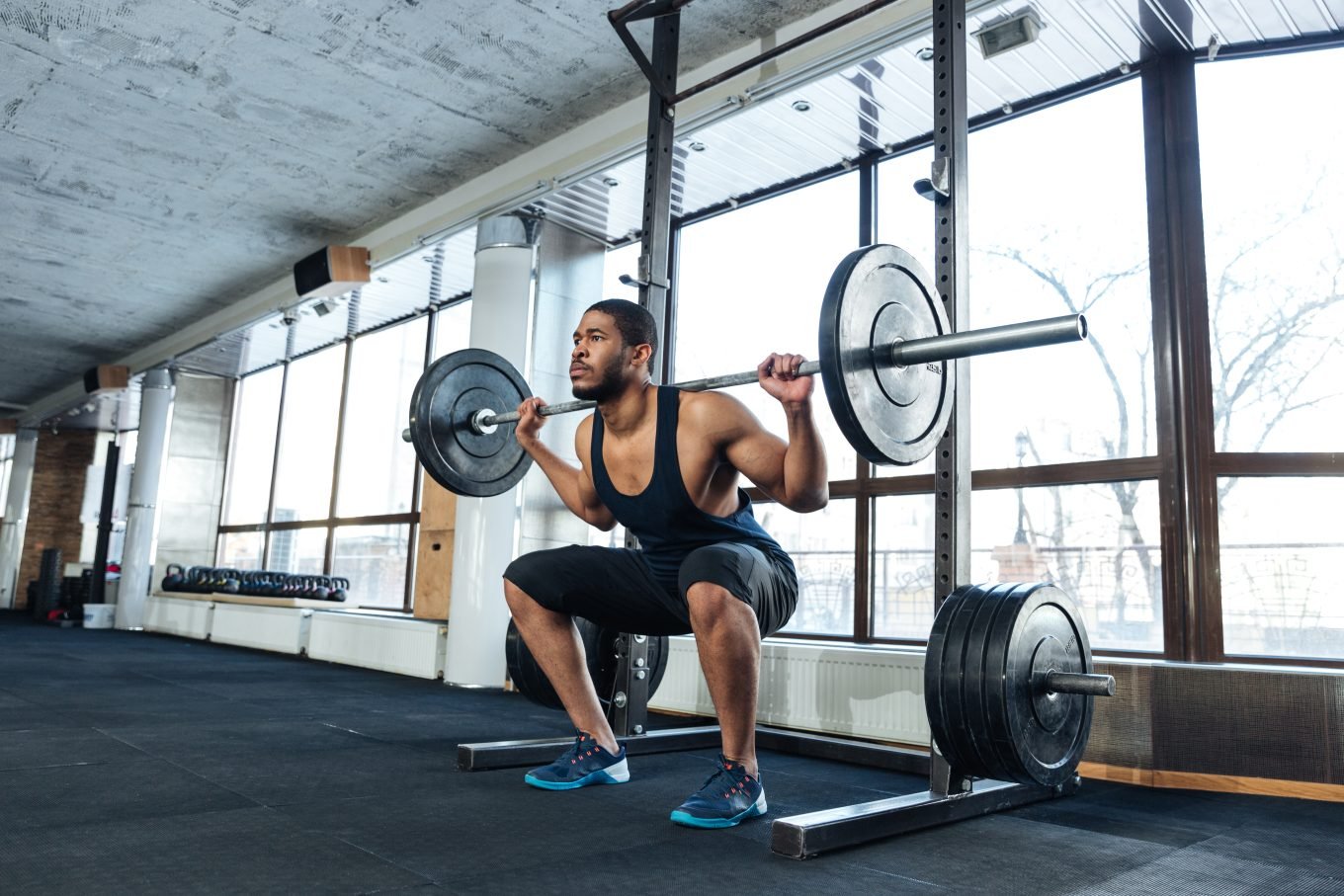 A black man performs a back barbell squat in front a squat rack in an airy gym. 