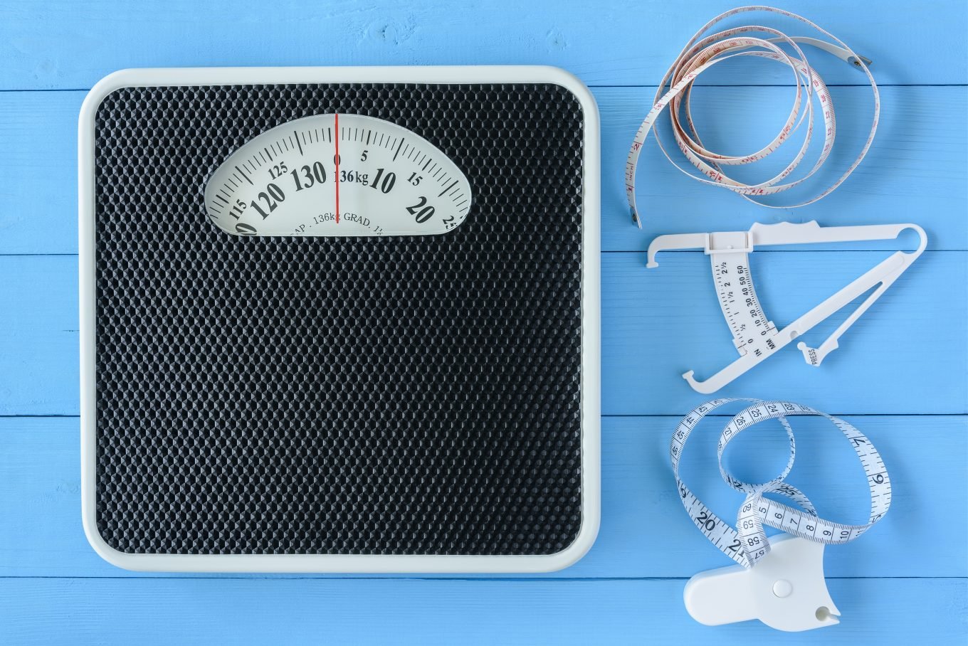 A textured black and white mechanical scale sits alongside a tape measure and skinfold calipers on a blue painted wooden background.