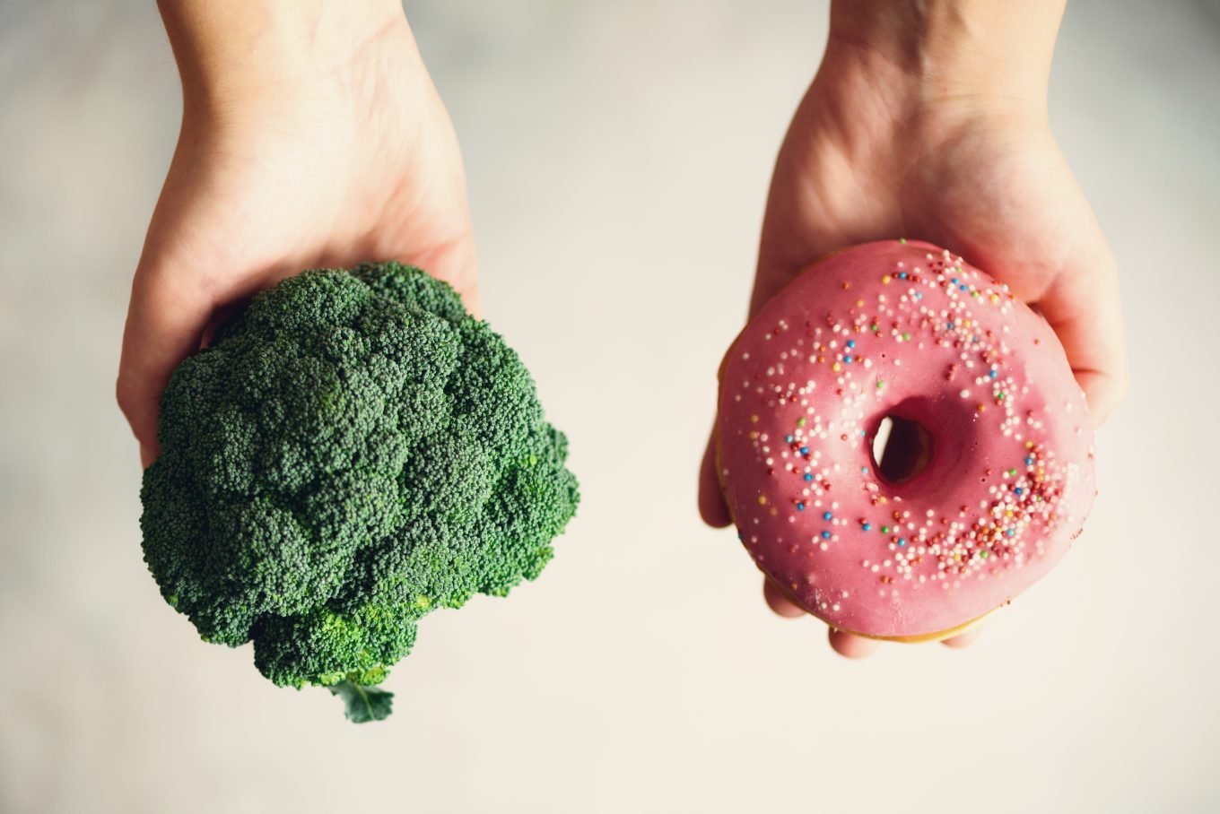 Broccoli and a pink donut are held in a person's palms, face up, as they choose which to eat. 