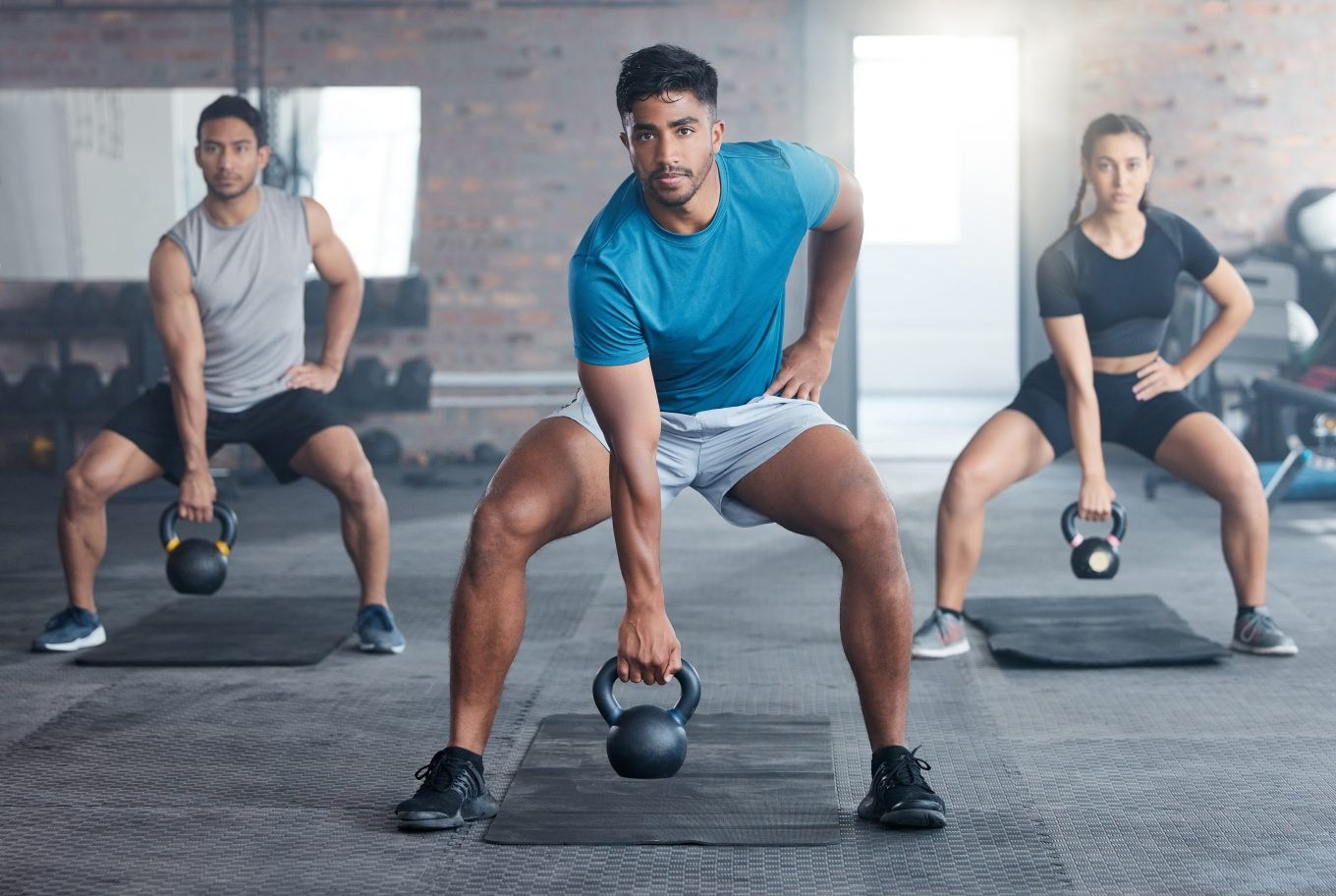 3 athletic young adults perform a kettlebell workout in a gym.