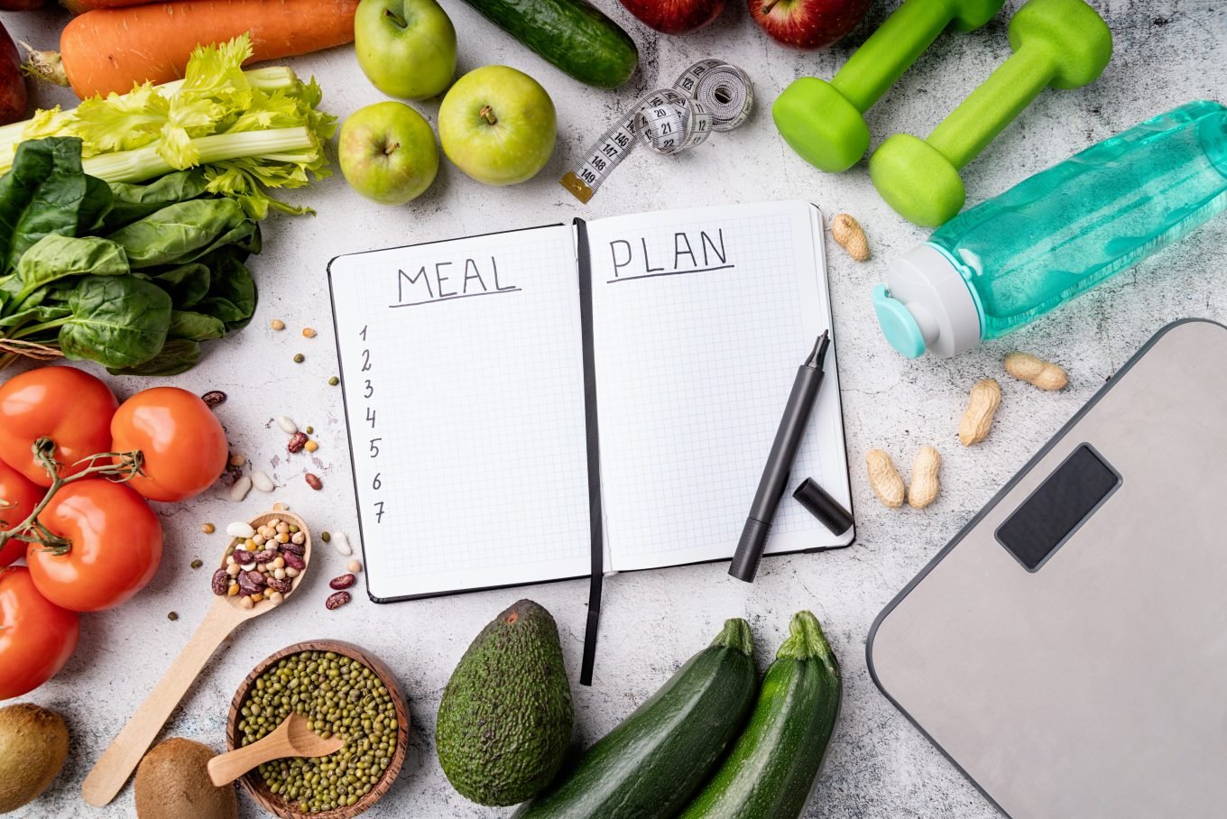 A note pad open to a page titled "Meal Plan" lays among a variety of healthy fruits and vegetables and fitness tools. 