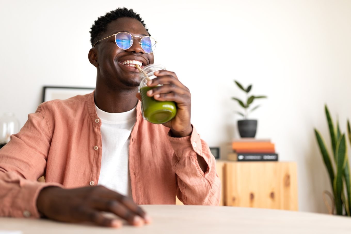 A young, black man grins while drinking a green juice from a mason jar through a straw. 