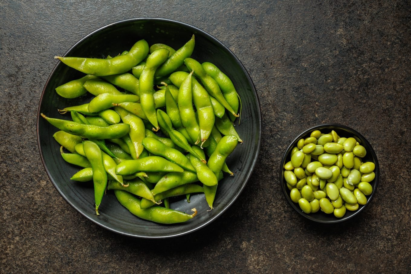 A large bowl of edamame pods sits next to a smaller bowl of edamame beans. 
