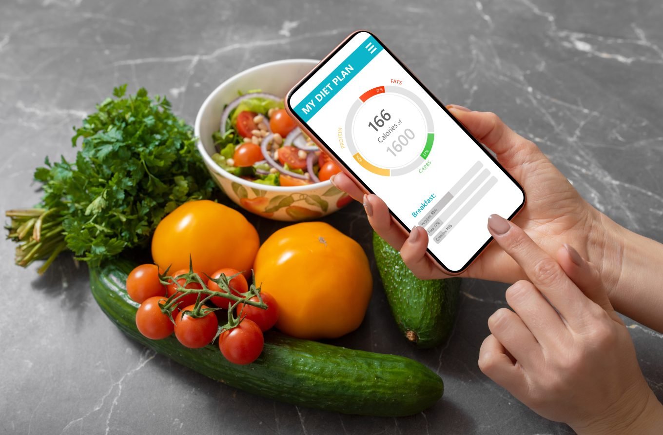 A smartphone displaying a calorie and macro counting app is held over a selection of fruits and vegetables. 