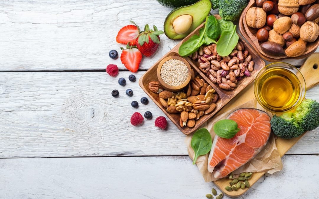 A selection of healthy foods arranged on a wooden surface, including a bowl of nuts, half an avocado, strawberries, and a cut of salmon.